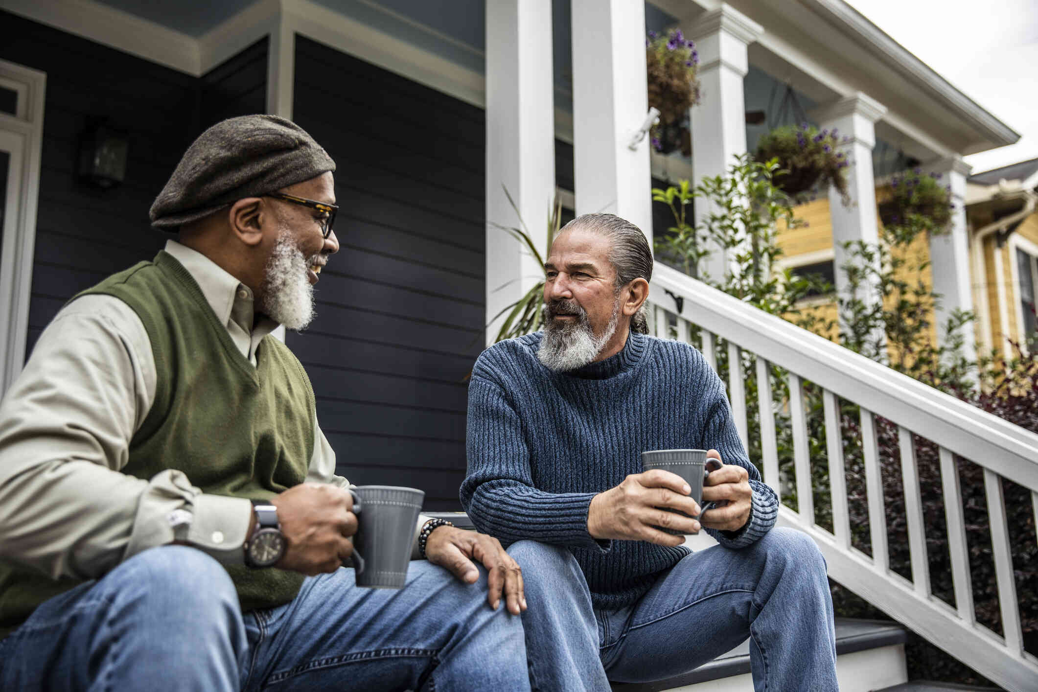 Two middle aged men sit side by side on the front porch steps and chat while smiling and holding coffee cups.